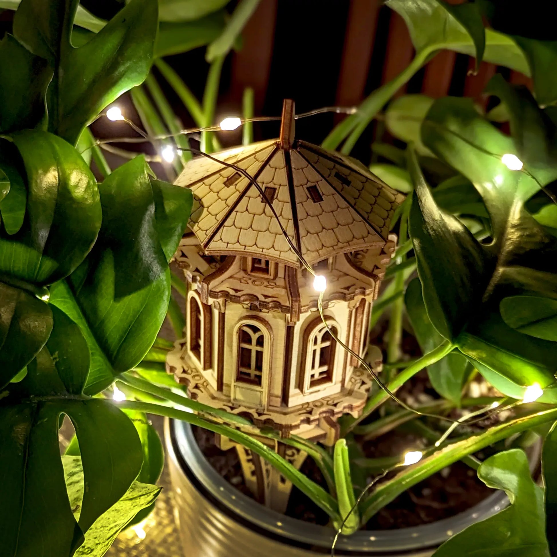 Decorative laser cut plywood garden tower staked in a plant pot, surrounded by lush leaves and glowing fairy lights.