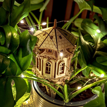 Decorative laser cut plywood garden tower staked in a plant pot, surrounded by lush leaves and glowing fairy lights.