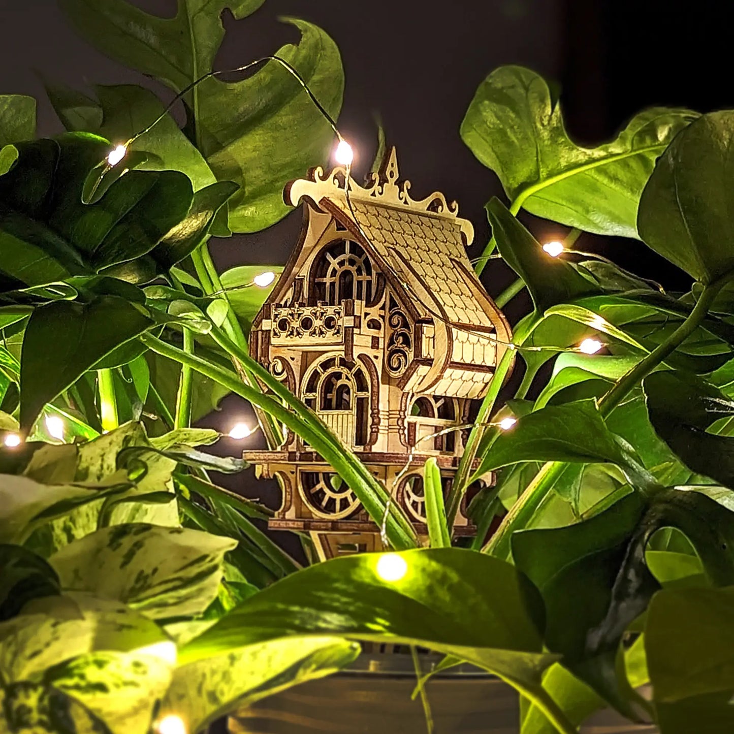 Magical laser cut plywood fairy house hidden inside lush green monstera leaves, illuminated by glowing warm led lights.