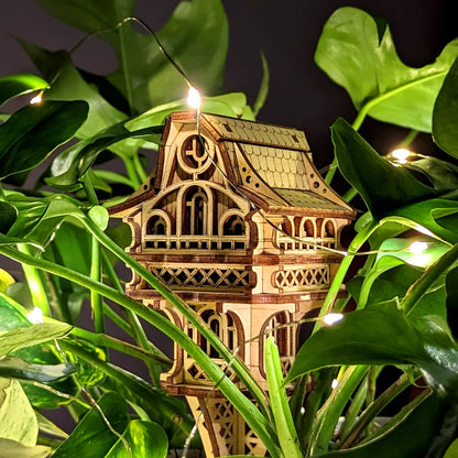 Small laser cut plywood gnome house nestled among green monstera leaves, illuminated by warm fairy lights for a magical look.