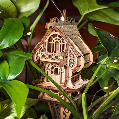 Fairy house made of laser cut plywood nestled among green plant leaves and warm fairy lights, creating cozy vibe.