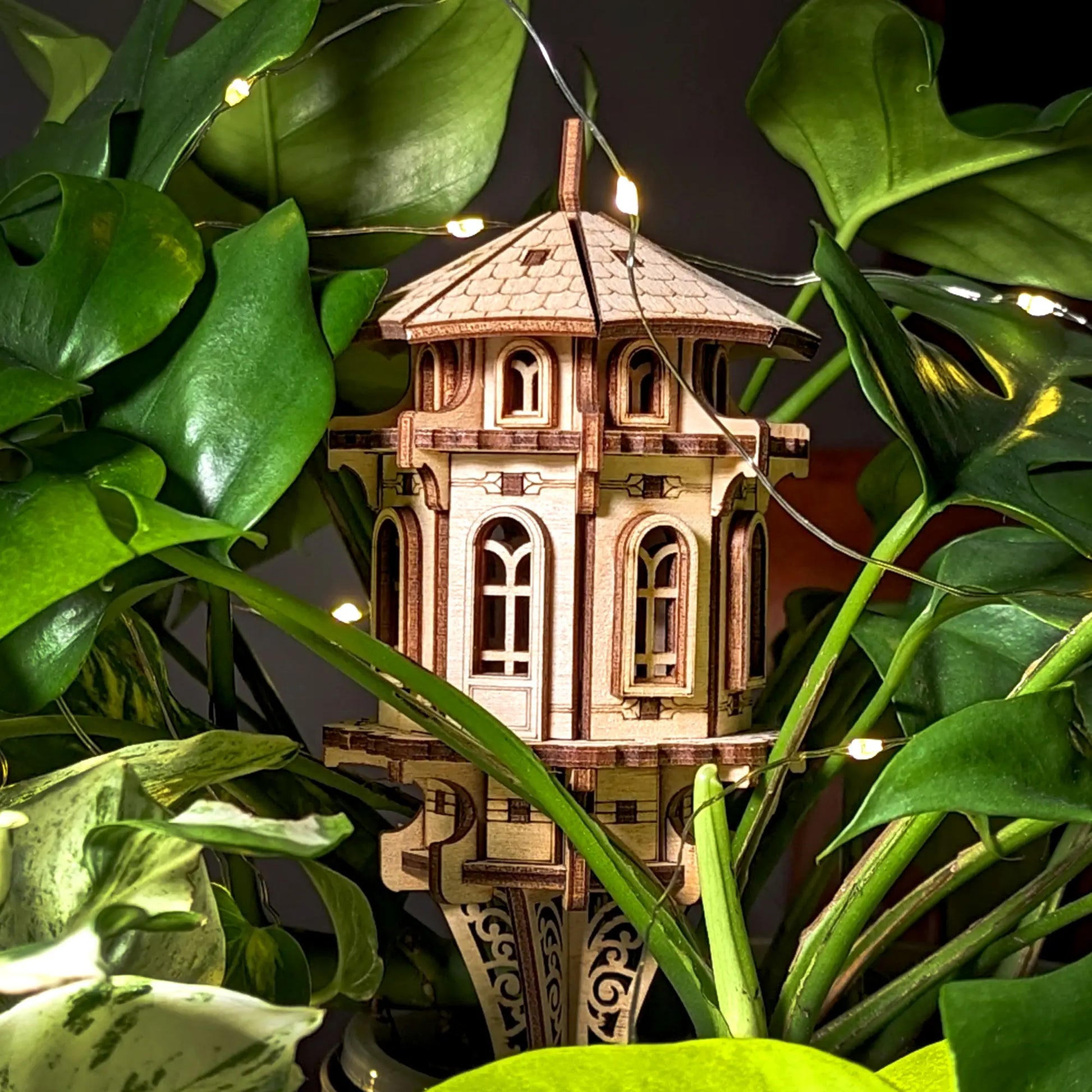 Laser cut plywood garden tower nestled in lush green indoor plants, accented by soft fairy lights for a magical view.