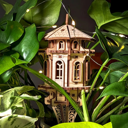 Laser cut plywood garden tower nestled in lush green indoor plants, accented by soft fairy lights for a magical view.