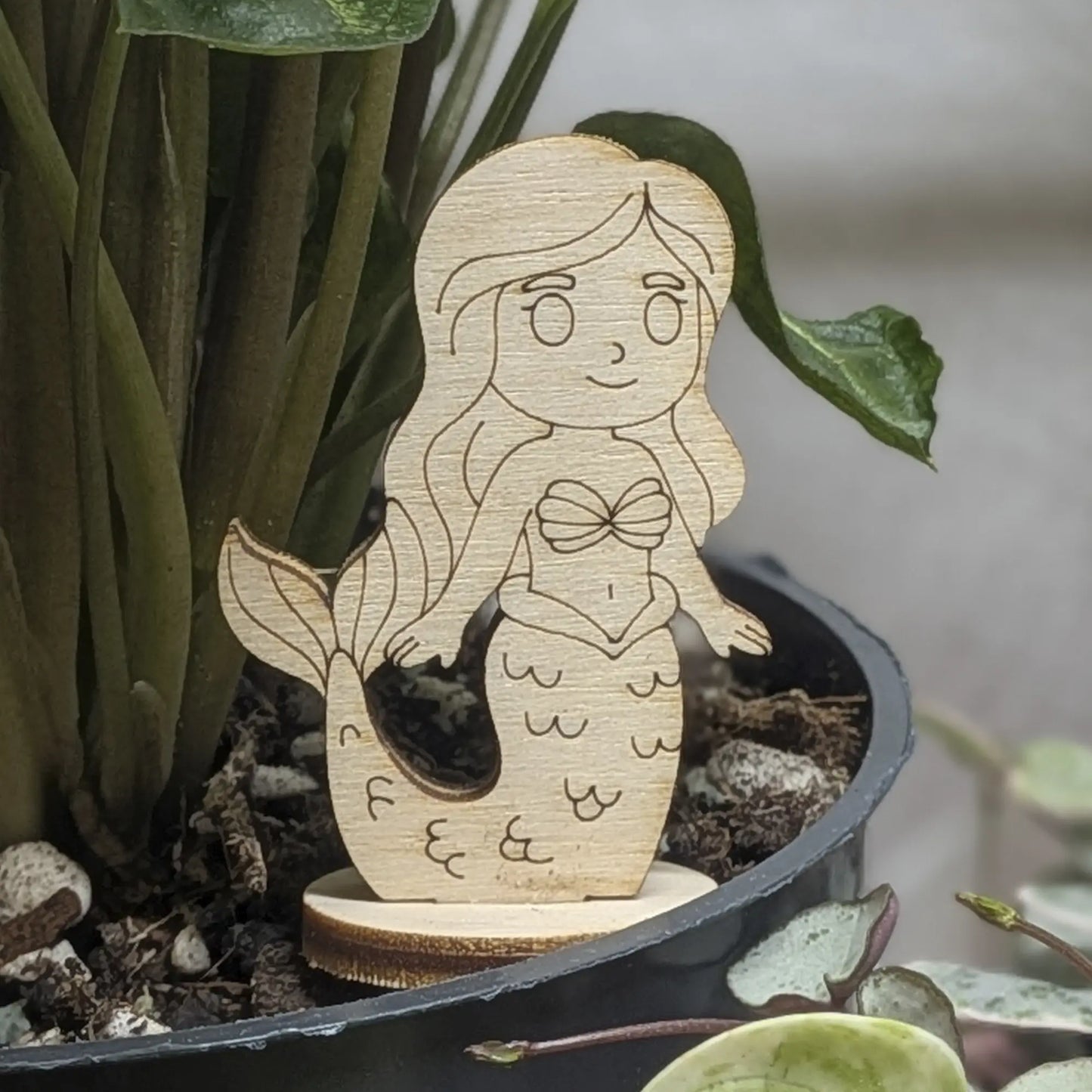 Laser cut plywood mermaid toy standing in a green plant pot. Detailed shell top, long hair, and scales engraved on wood