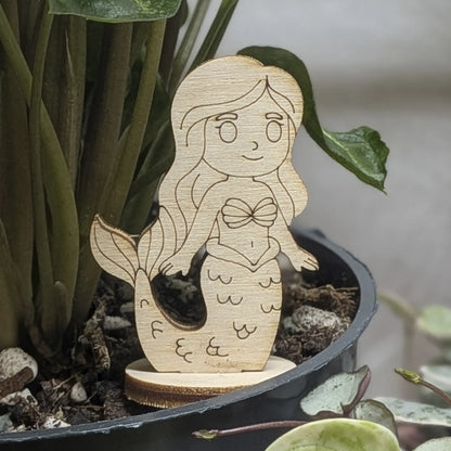 Laser cut plywood mermaid toy standing in a green plant pot. Detailed shell top, long hair, and scales engraved on wood