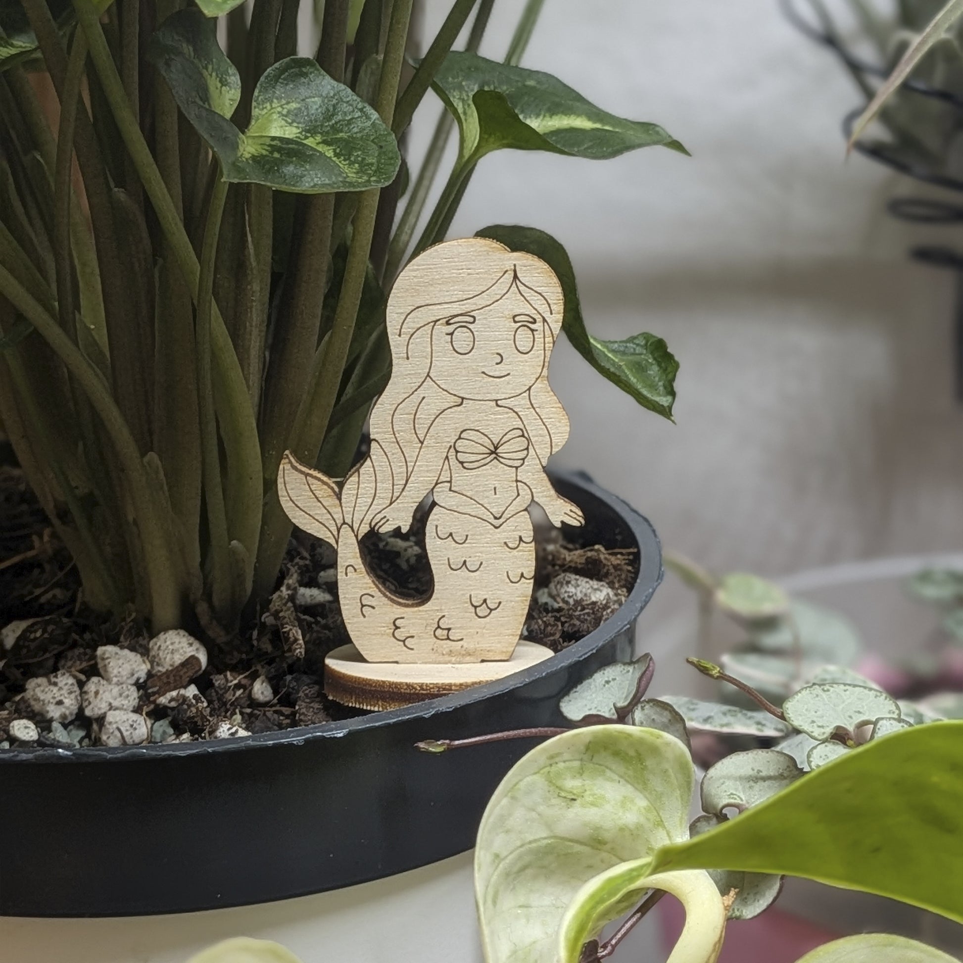 Laser cut plywood mermaid toy standing in a green plant pot. Detailed shell top, long hair, and scales engraved on wood