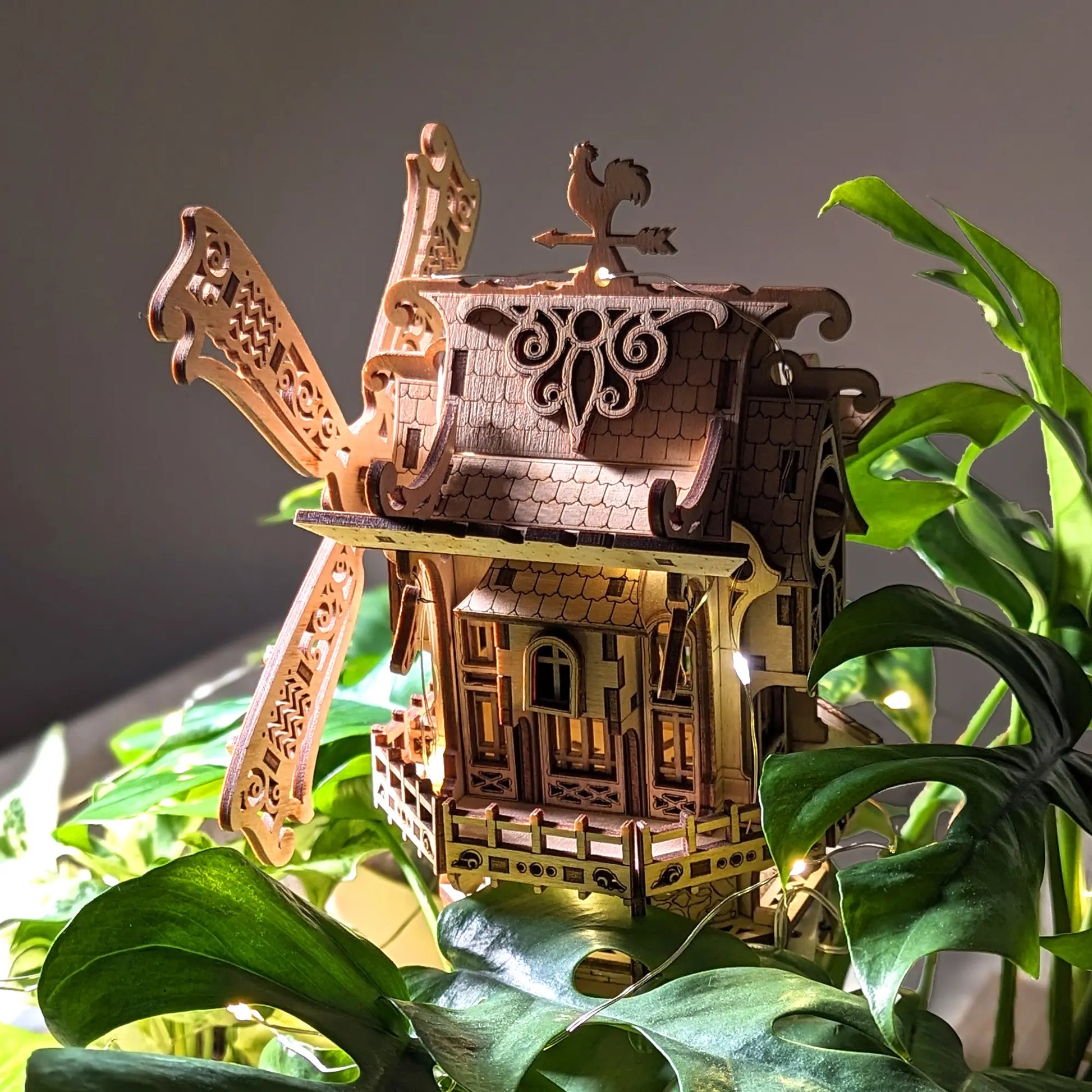 An ornate wooden village windmill with a rooster weather vane nestled in lush green plant leaves, made of laser cut plywood.