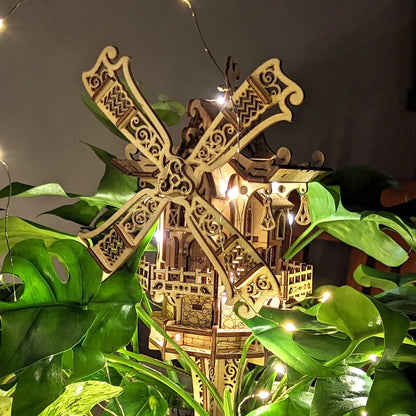 An atmospheric cozy shot of a glowing 3D miniature windmill hidden in deep green Monstera leaves, made of laser cut plywood.