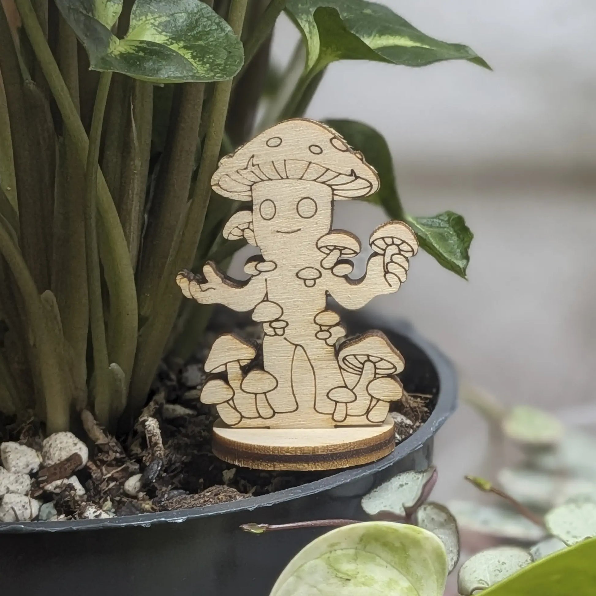 Laser cut plywood mushroom folk figurine sits in a green plant pot. Features a spotted cap, small arms, and solid wood base
