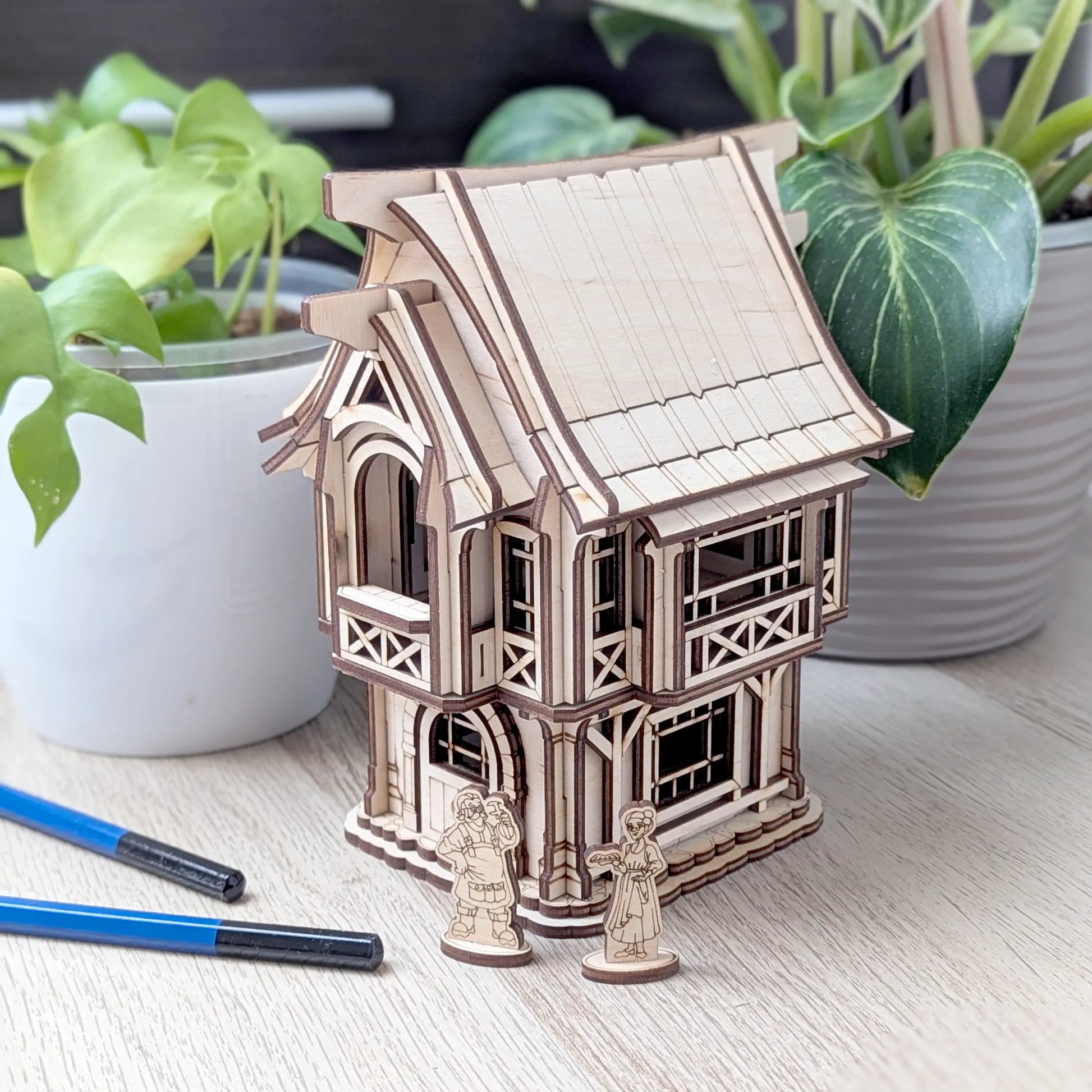 A beautiful wide view of an assembled rustic laser cut plywood old village house on a table with green potted house plants.