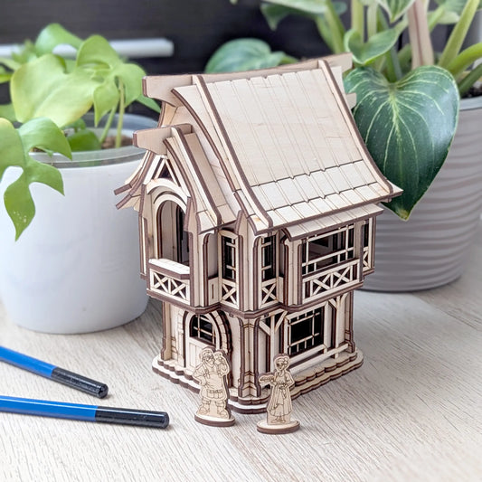 A beautiful wide view of an assembled rustic laser cut plywood old village house on a table with green potted house plants.