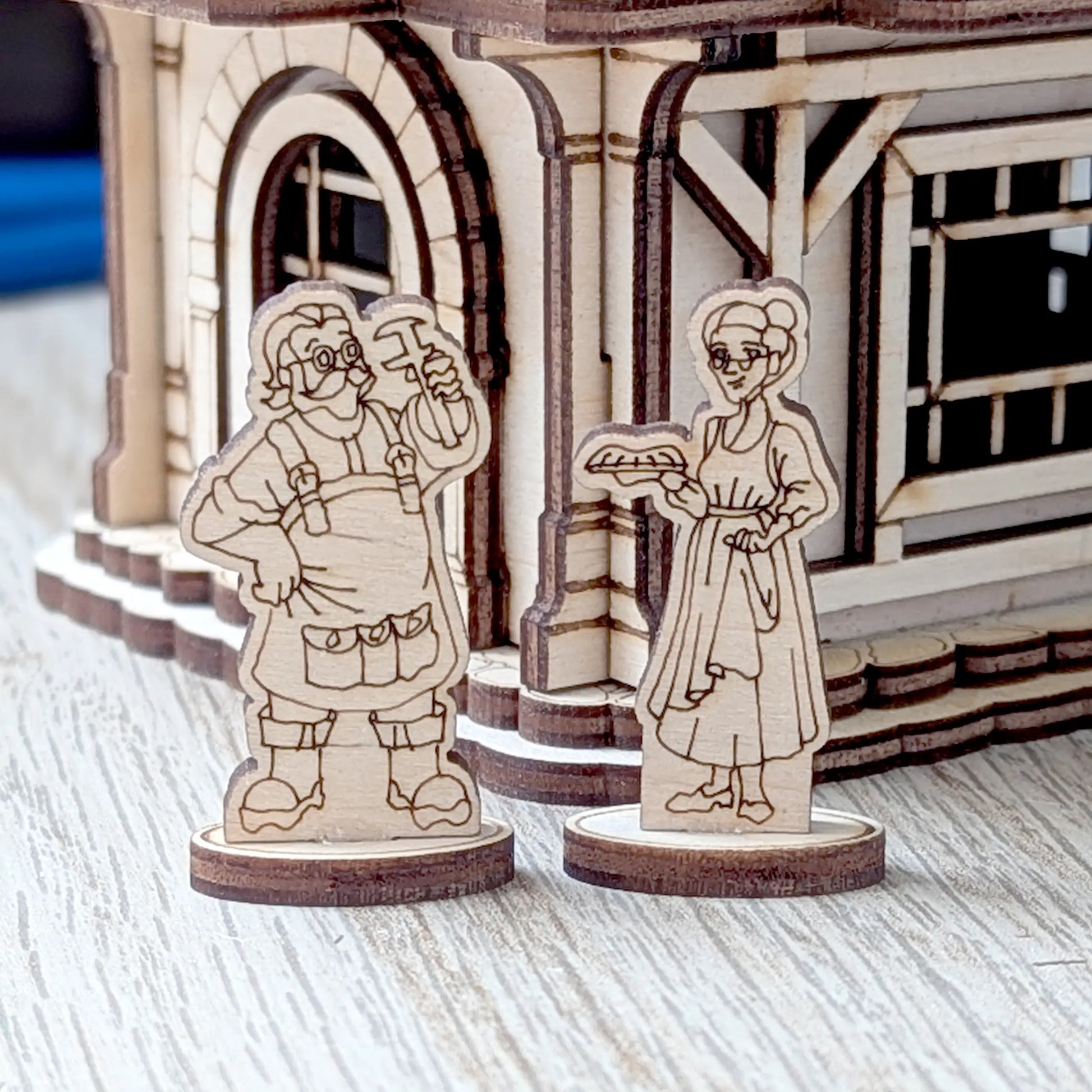A close-up of two detailed miniature wooden villager figurines standing outside the door of a laser cut plywood dollhouse.