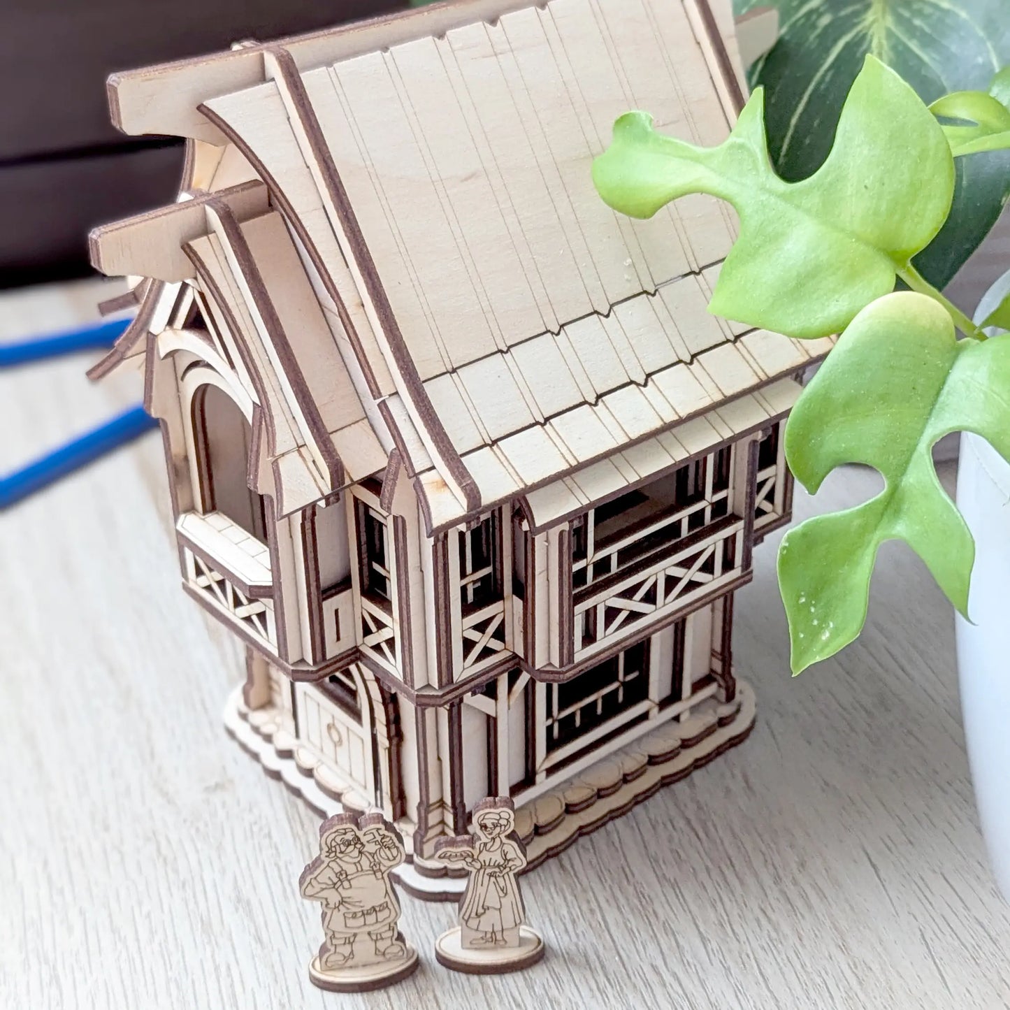 An aesthetic lifestyle shot of a miniature rustic old village house on a round woven mat surrounded by green houseplants.