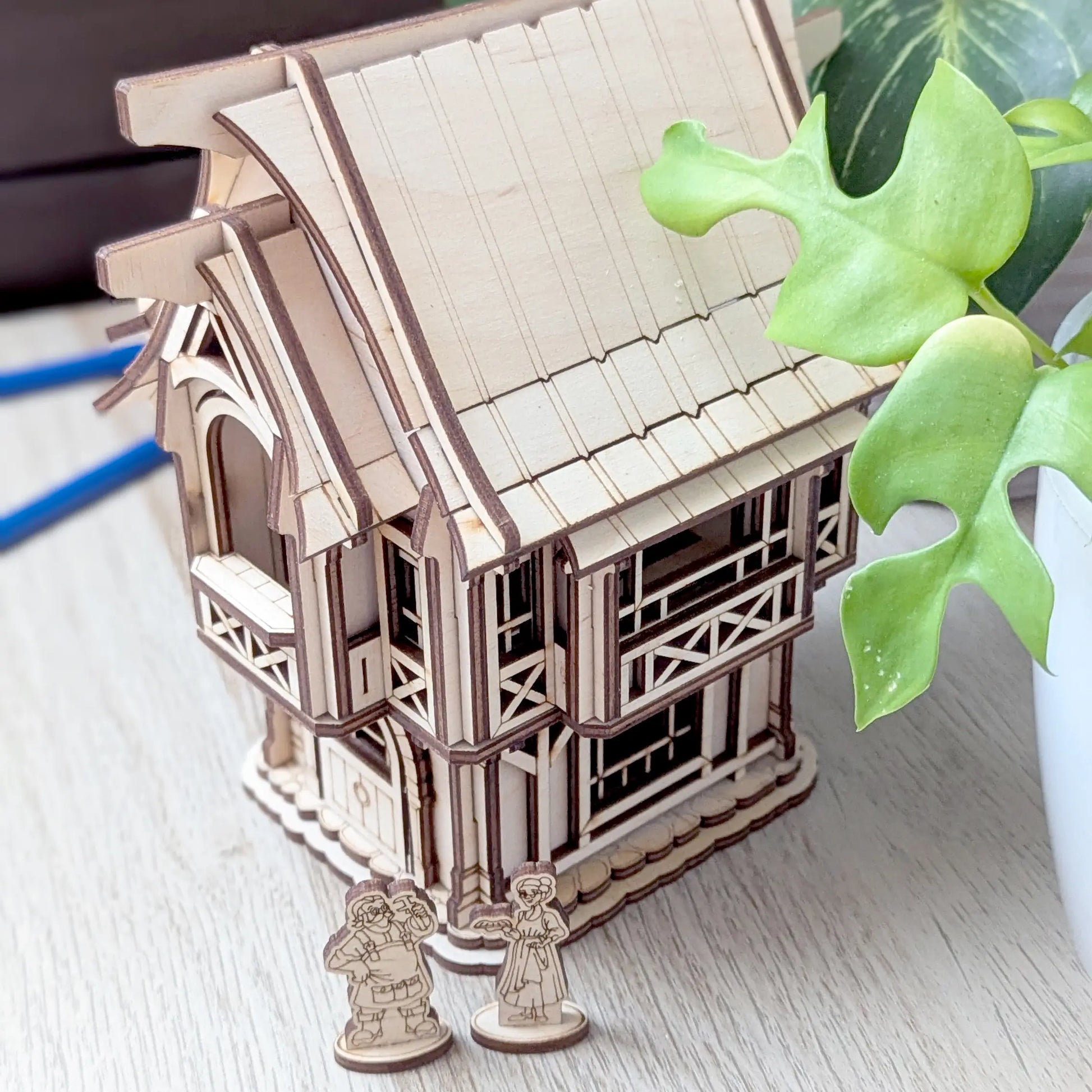An aesthetic lifestyle shot of a miniature rustic old village house on a round woven mat surrounded by green houseplants.