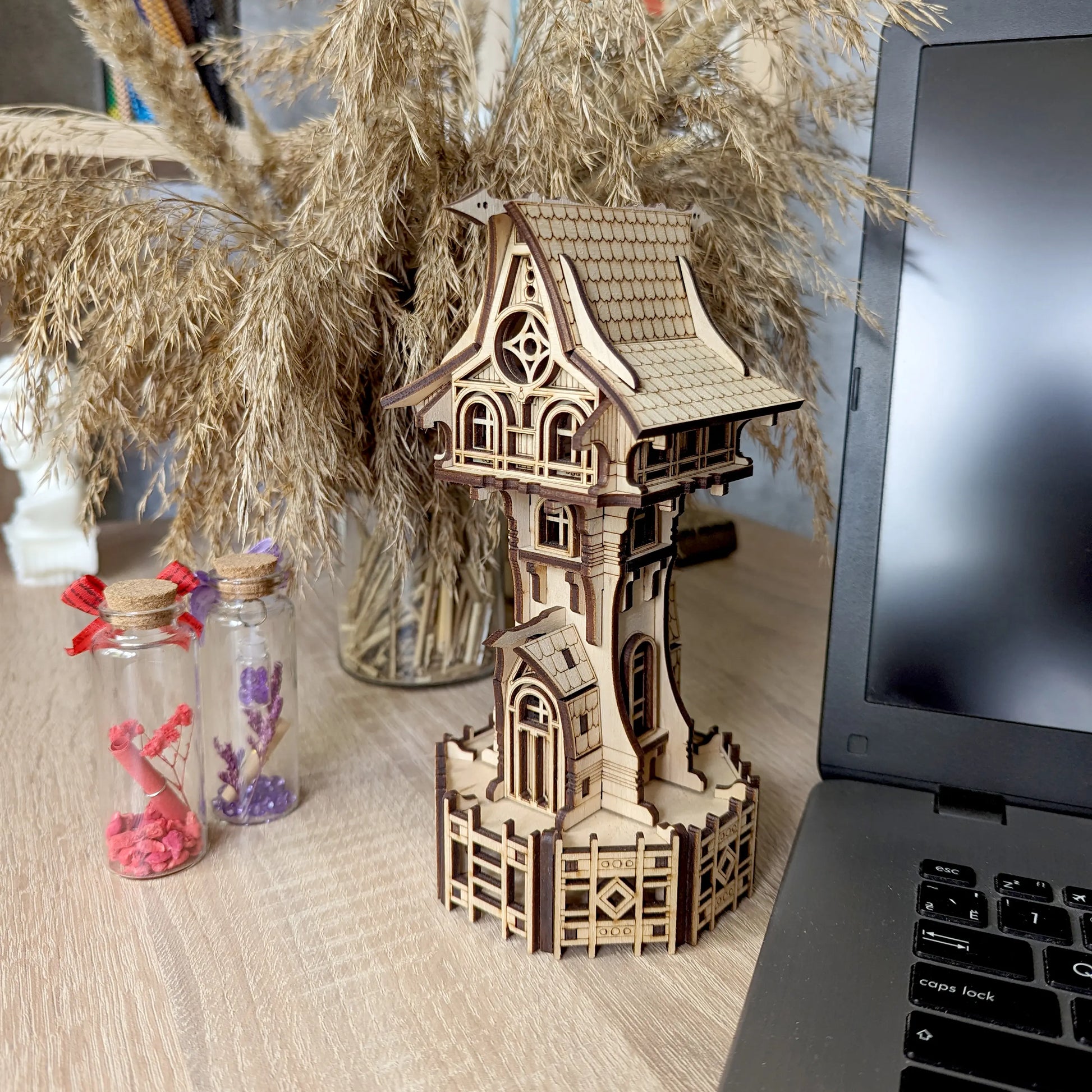 Laser cut plywood miniature tower sits on a table near a laptop, dried grass bouquet, and glass bottles.