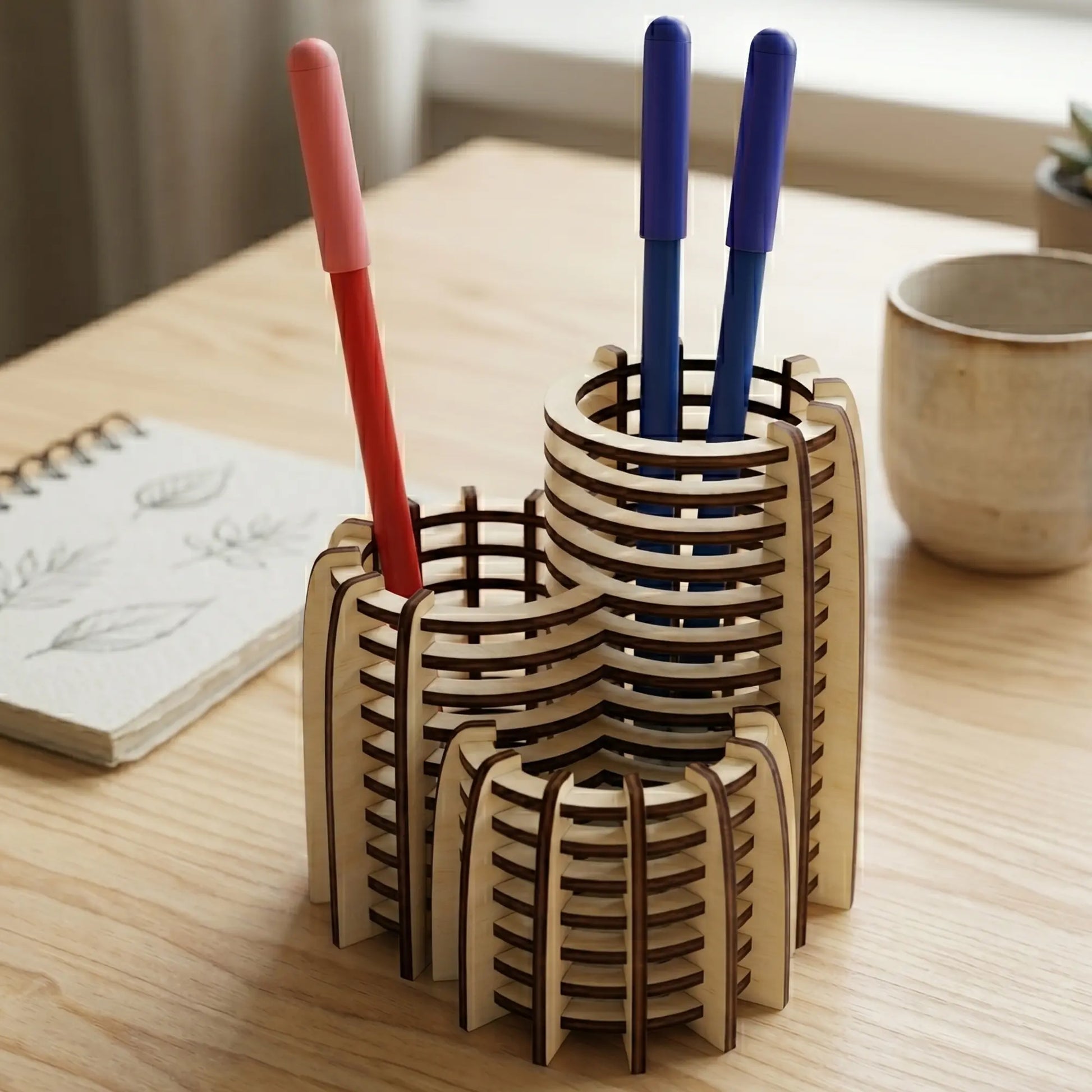 Modern tiered laser cut plywood pen holder on a wooden desk with a notebook and pens in a bright, sunlit Scandinavian room.