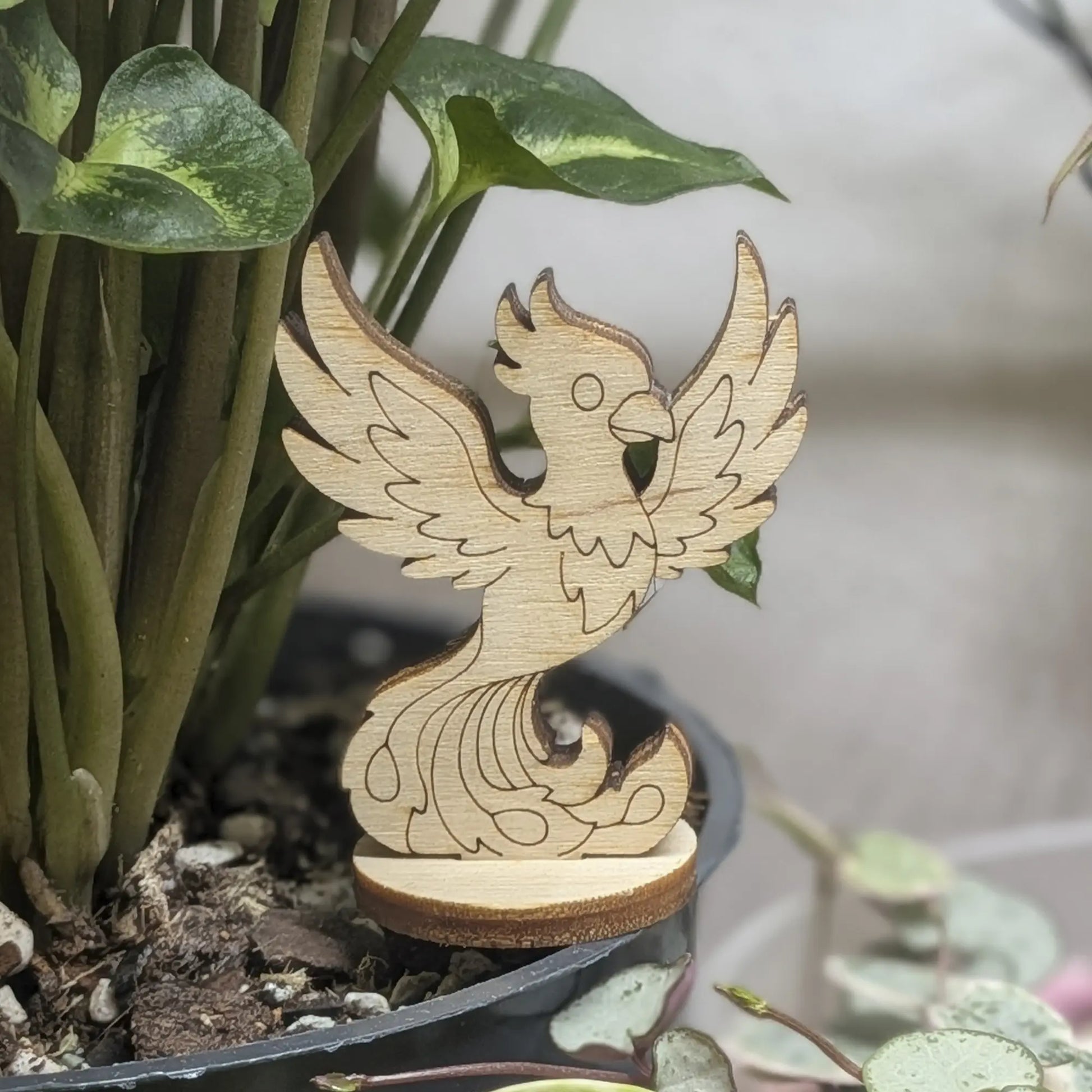 A laser cut plywood phoenix figurine stands in a plant pot. Features engraved wings, crest, and flaming tail on wood base