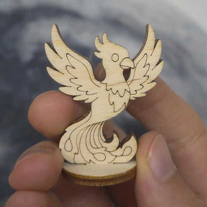 Hand holds a laser cut plywood phoenix figurine. Features detailed engraved feathers, sharp beak, and flaming tail plumes