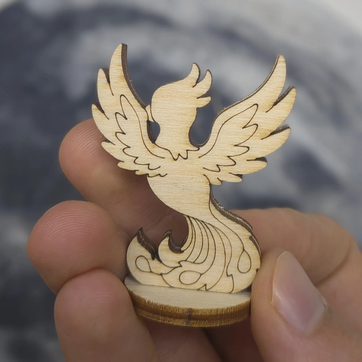 Back view of laser cut plywood phoenix held in hand. Shows dark burnt edges on base and intricate wing feather engravings