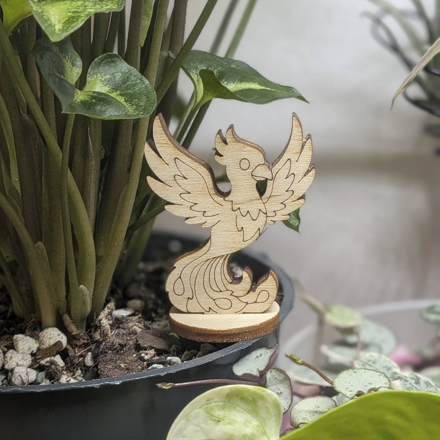 A laser cut plywood phoenix figurine stands in a plant pot. Features engraved wings, crest, and flaming tail on wood base