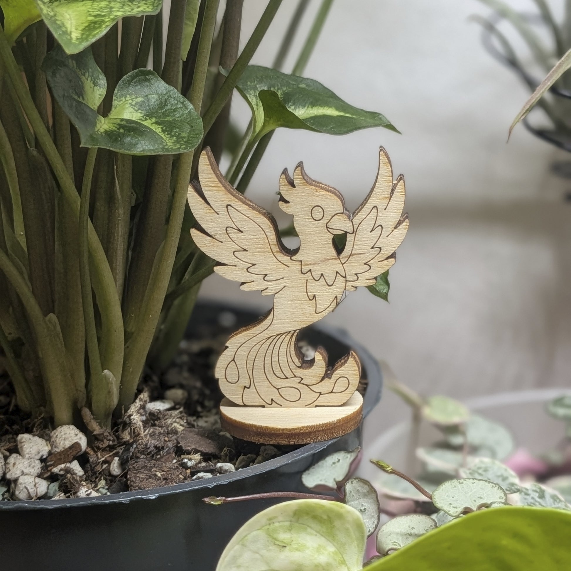 A laser cut plywood phoenix figurine stands in a plant pot. Features engraved wings, crest, and flaming tail on wood base