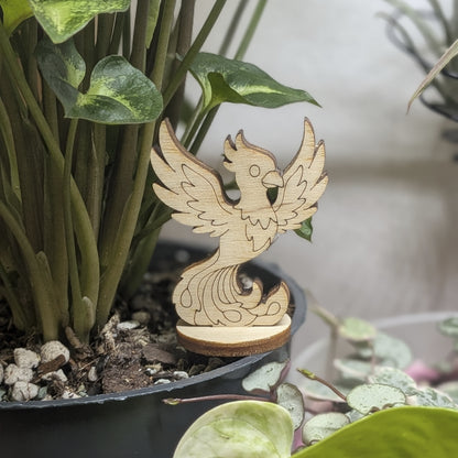 A laser cut plywood phoenix figurine stands in a plant pot. Features engraved wings, crest, and flaming tail on wood base