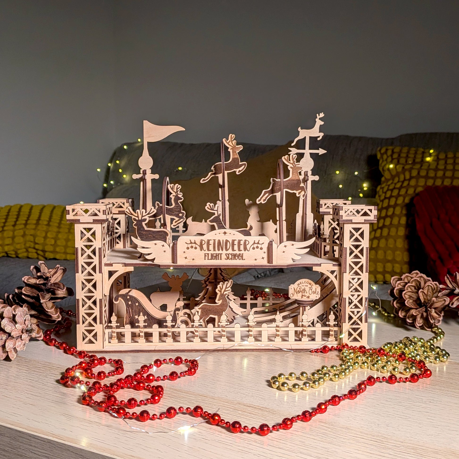 Assembled wooden “Reindeer Flight School” holiday decoration made from a laser cut digital file, shown on a table with festive lights, pinecones and beads. Layered reindeer shapes and towers create a detailed 3D scene
