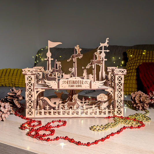 Assembled wooden “Reindeer Flight School” holiday decoration made from a laser cut digital file, shown on a table with festive lights, pinecones and beads. Layered reindeer shapes and towers create a detailed 3D scene