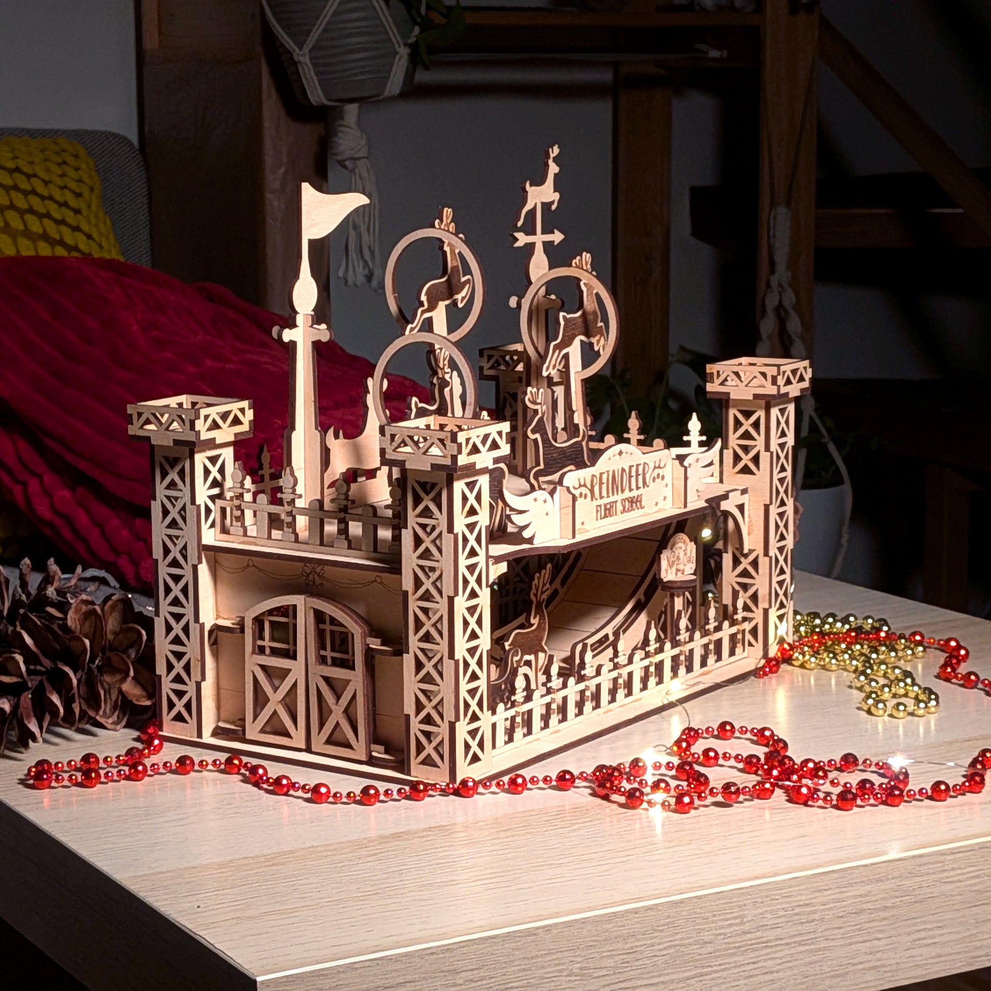 Wooden “Reindeer Flight School” holiday scene made from a laser cut digital file, displayed on a decorated table with pinecones, gold and red beads, and seasonal figurines