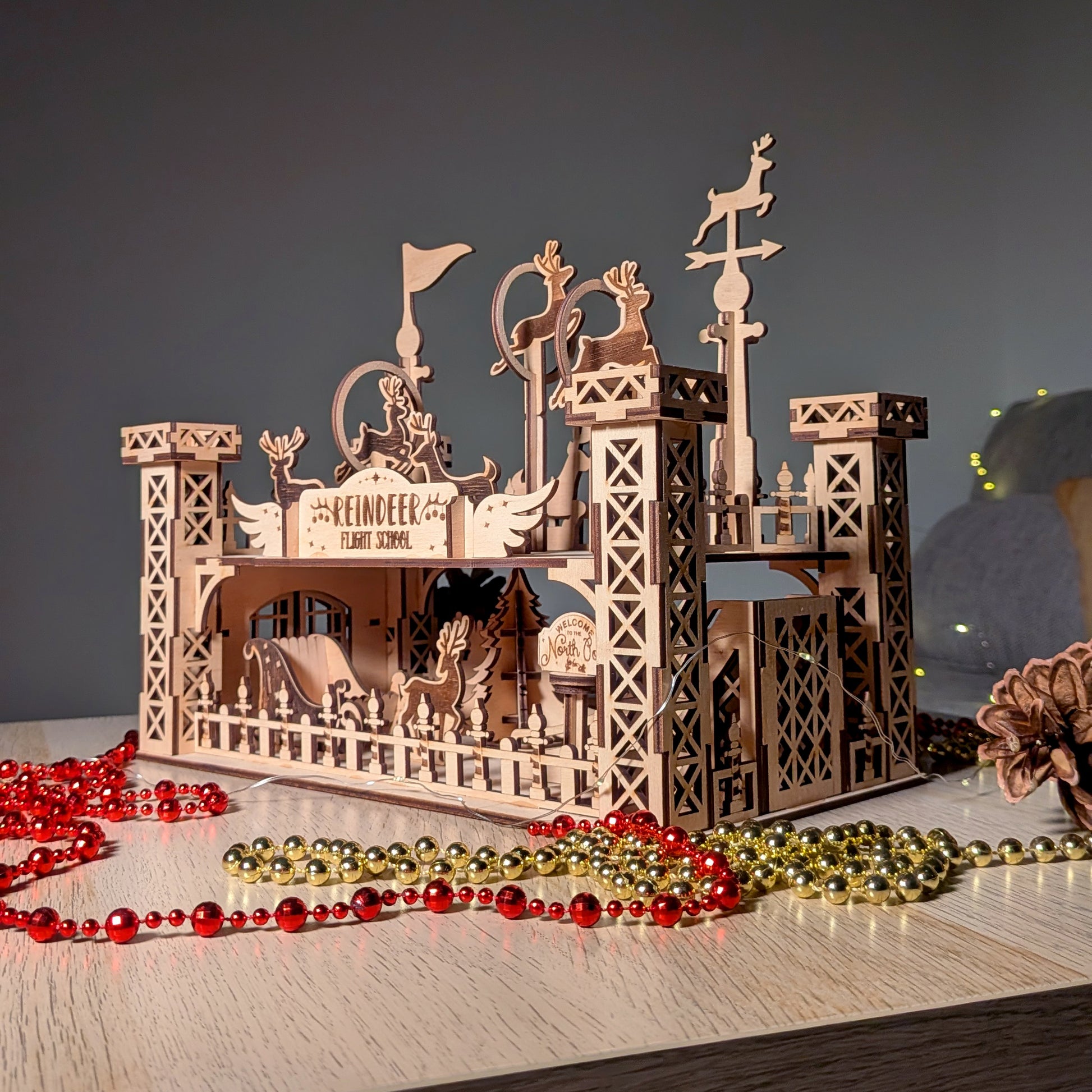 Wooden “Reindeer Flight School” display made from a laser cut digital design, featuring layered reindeer silhouettes on raised posts and a sleigh scene below, styled with pinecones and festive lights