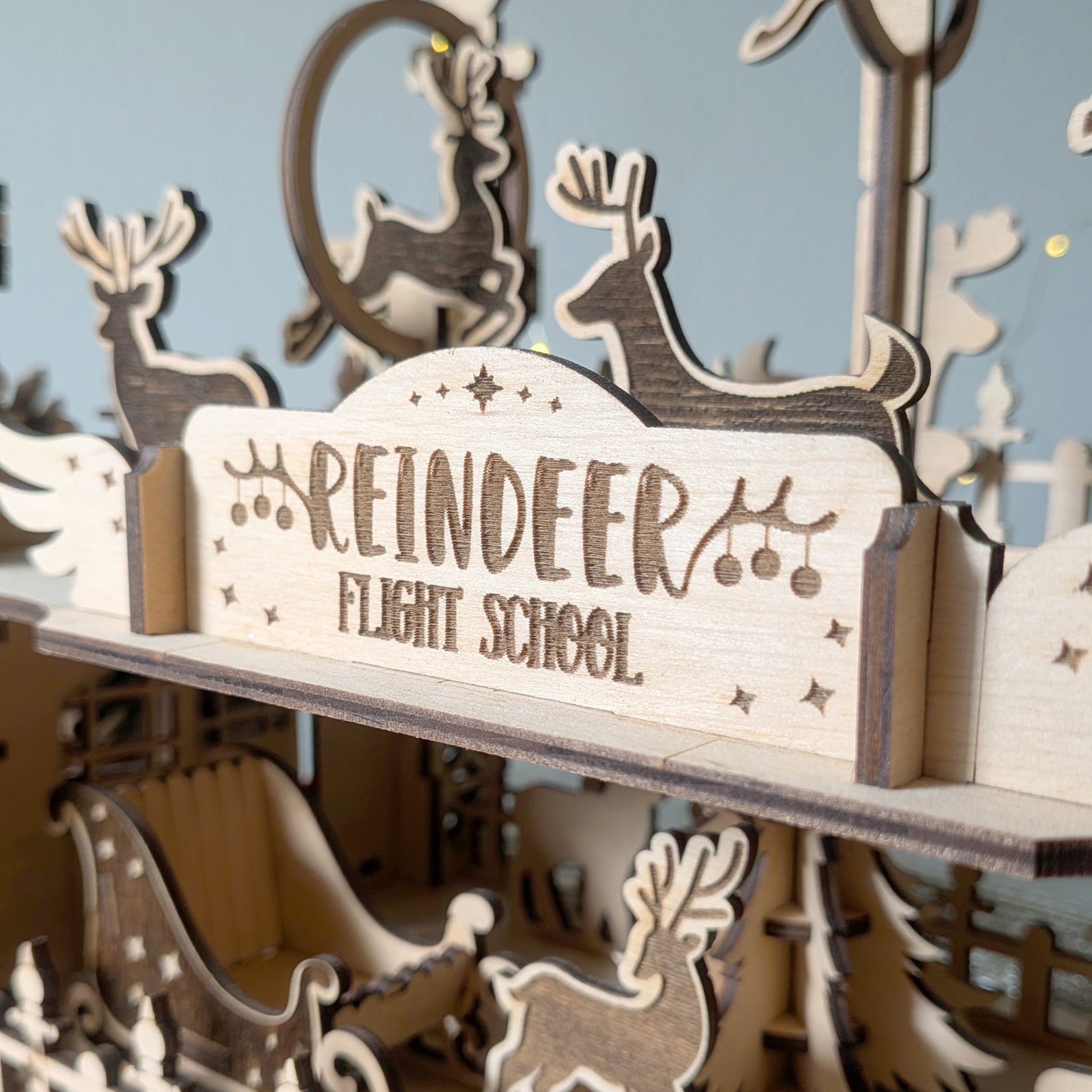 Close-up of the engraved “Reindeer Flight School” sign on a wooden holiday scene made from a laser cut digital file, showing detailed reindeer shapes and a sleigh in layered 3D construction.