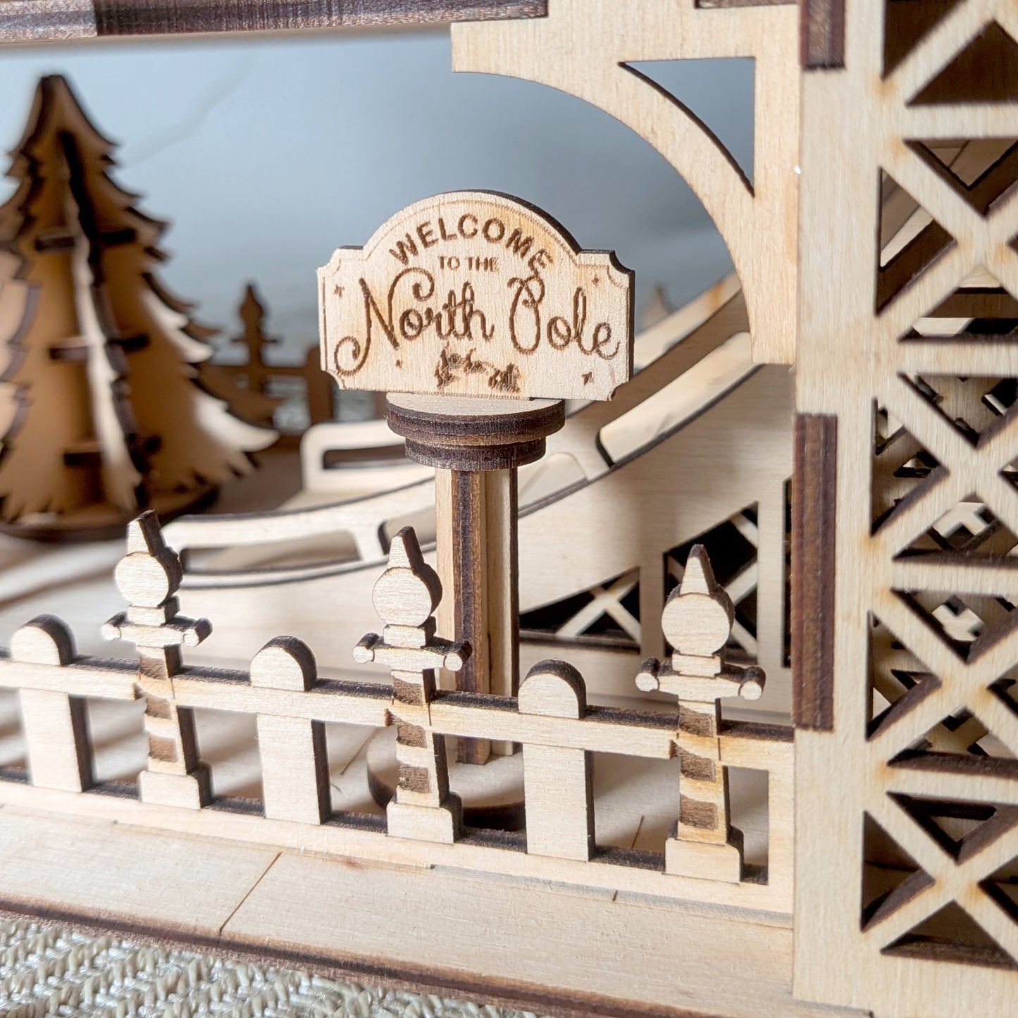 Close-up of the engraved “Welcome to the North Pole” sign in a wooden holiday scene made from a laser cut digital file, with fence and tree details in the background