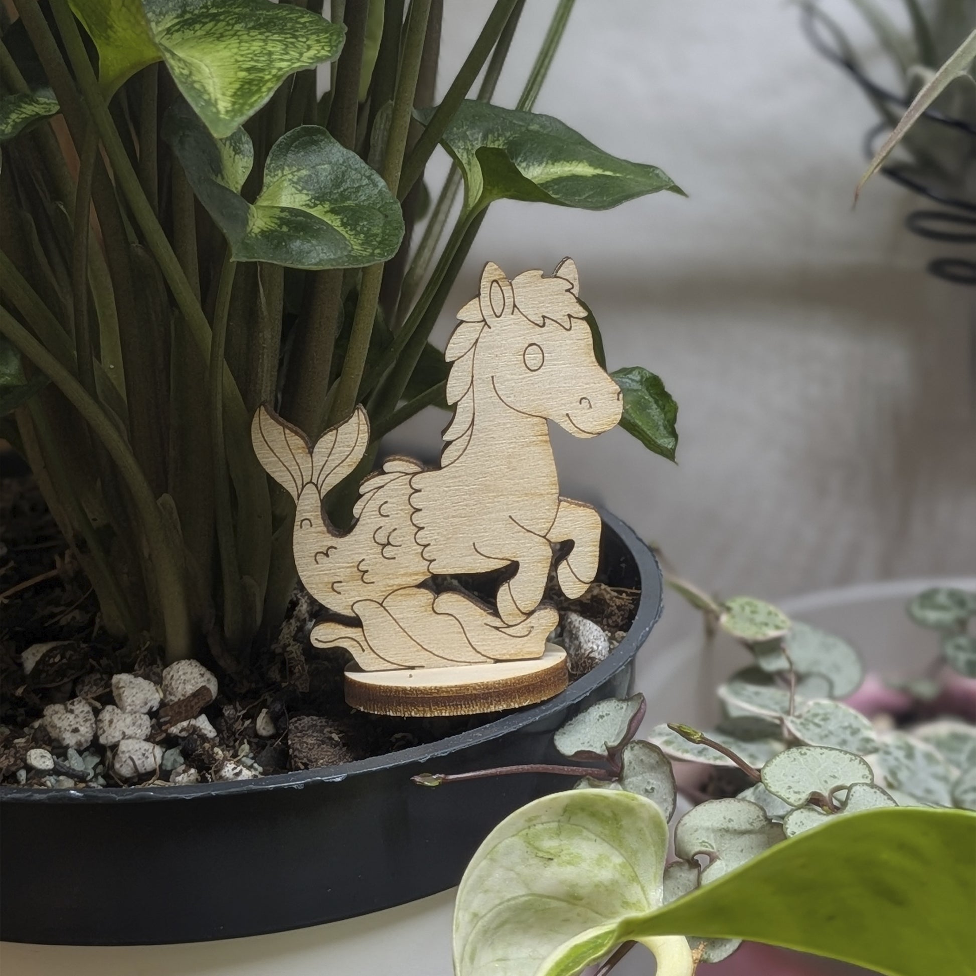 A wooden laser cut plywood seahorse mermaid  figurine is sitting in a potted plant, surrounded by green leaves and rocks.