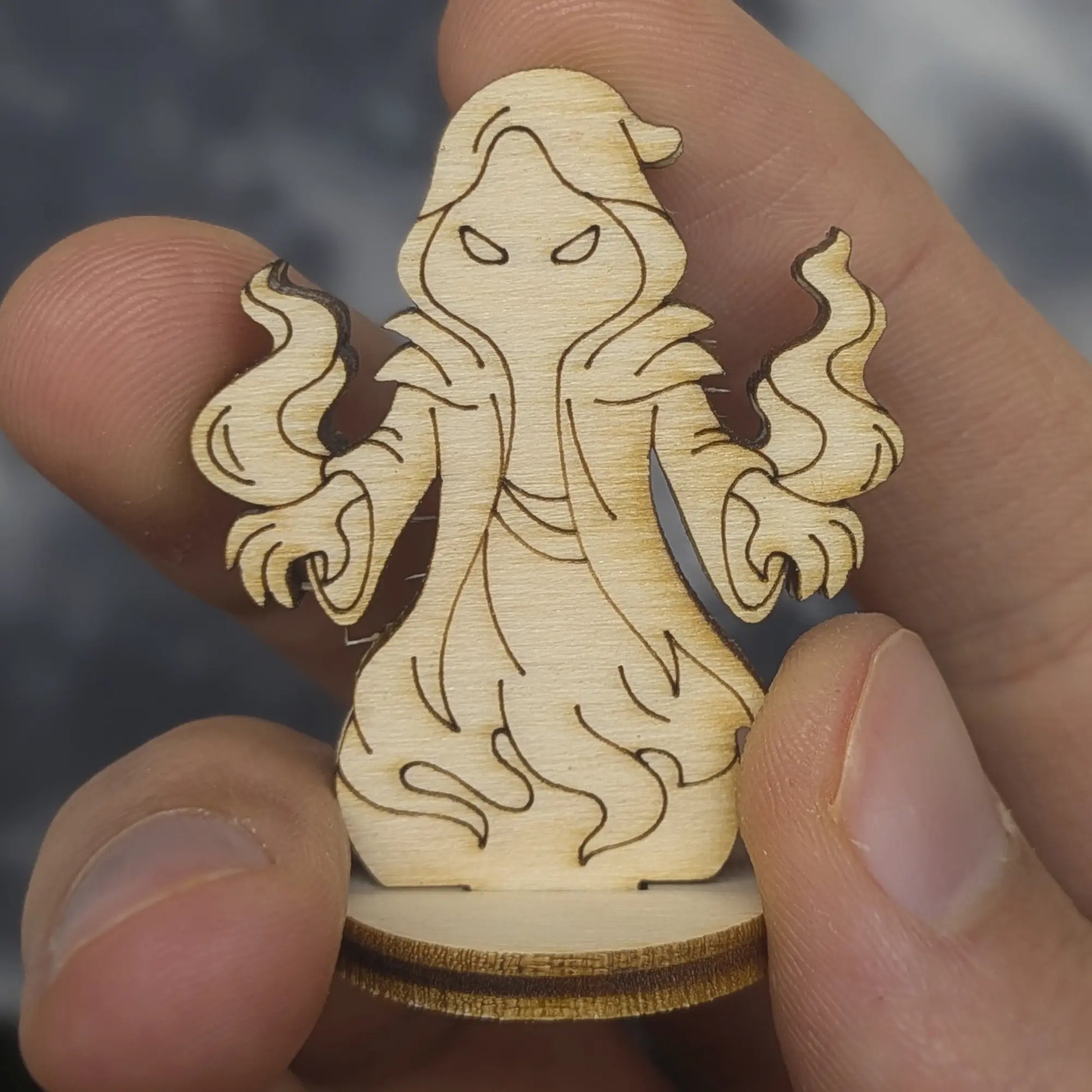 Hand holds a laser cut plywood shadow mage figurine. Shows a hooded robe, swirling magic, and dark burnt edges for detail