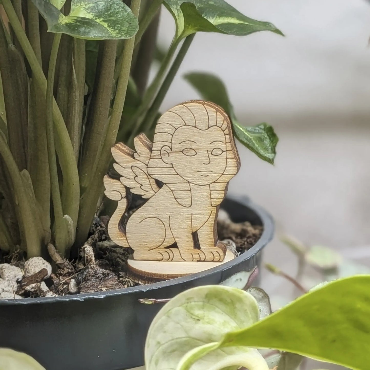 Winged sphinx figurine made of laser cut plywood sits in a flower pot. Features a human face, lion body, and fine engravings
