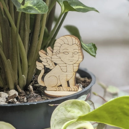 Winged sphinx figurine made of laser cut plywood sits in a flower pot. Features a human face, lion body, and fine engravings