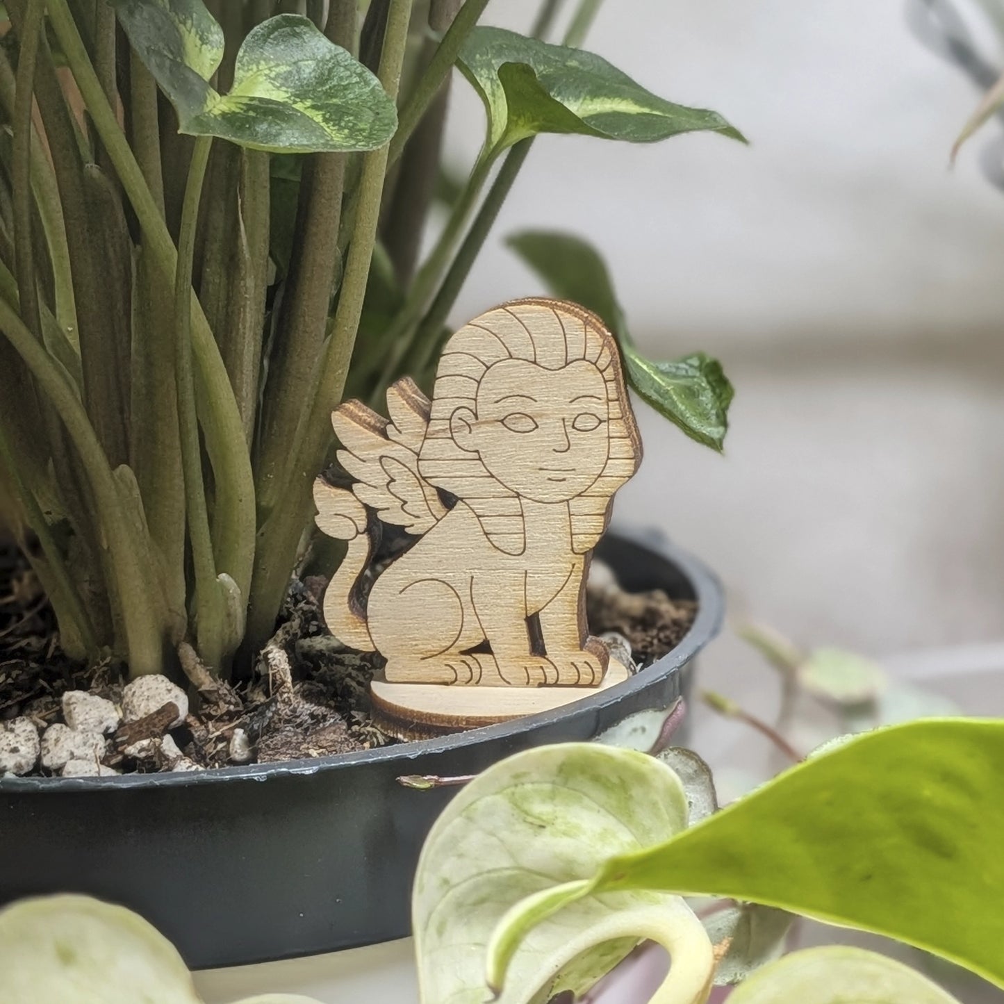 Winged sphinx figurine made of laser cut plywood sits in a flower pot. Features a human face, lion body, and fine engravings