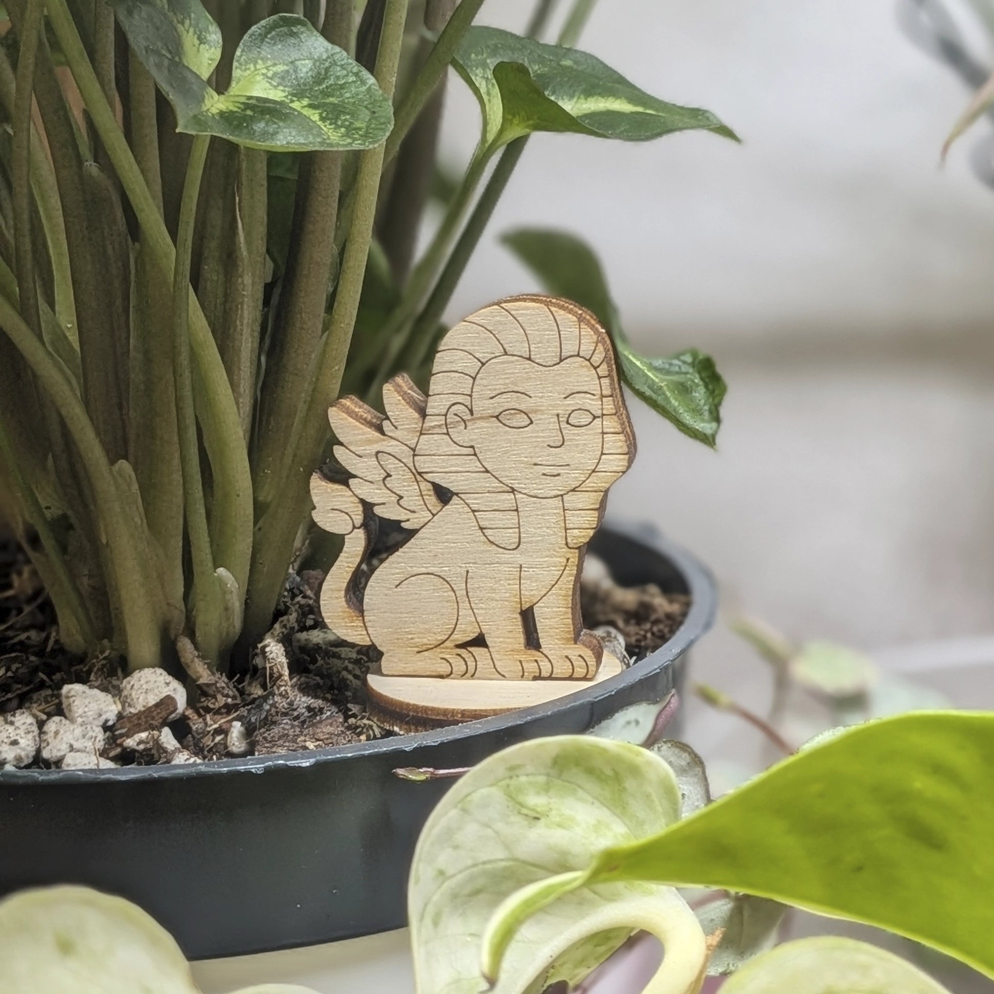 Winged sphinx figurine made of laser cut plywood sits in a flower pot. Features a human face, lion body, and fine engravings