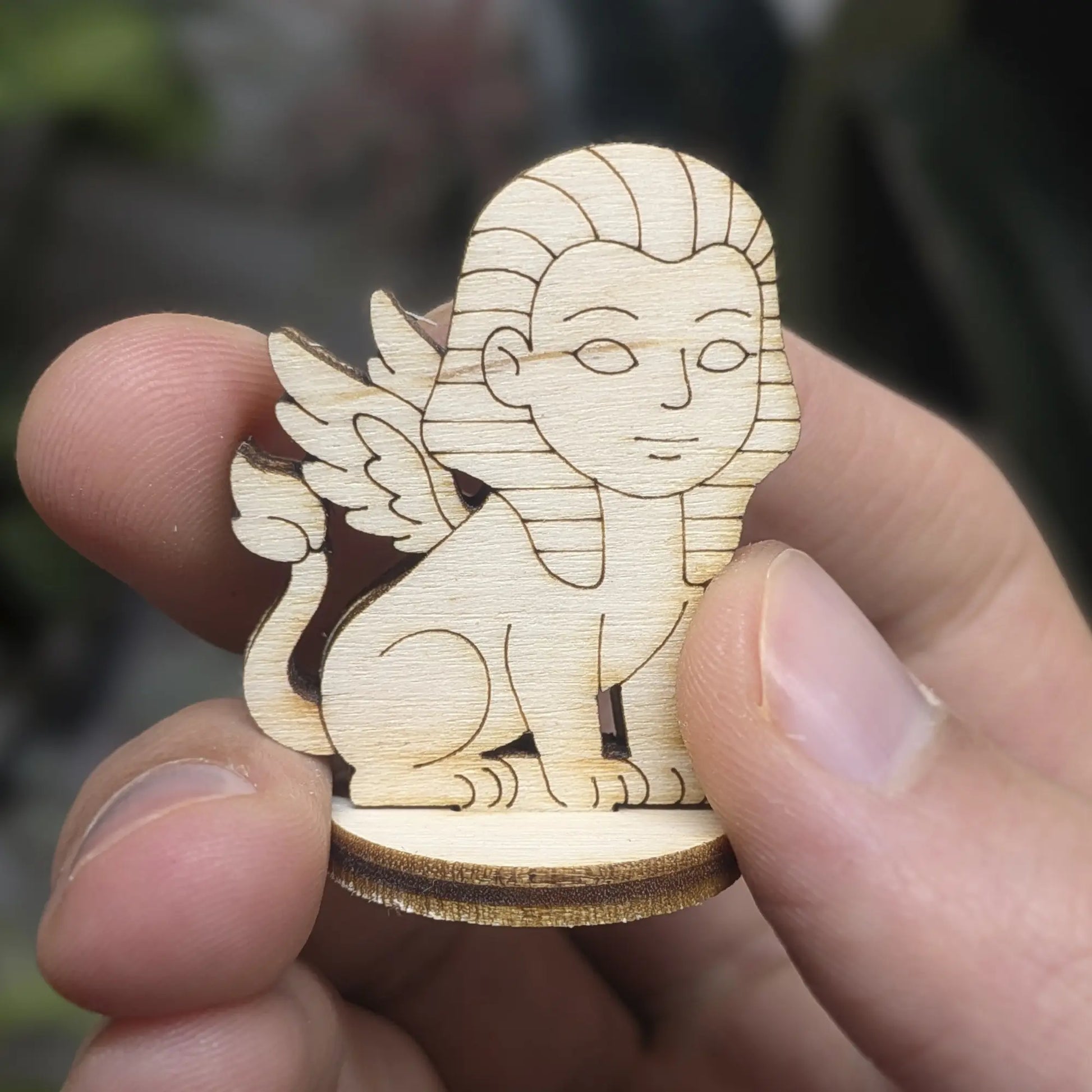 Hand holds a mini sphinx of laser cut plywood. Showing pharaonic headpiece, wings, and engraved details at a small scale