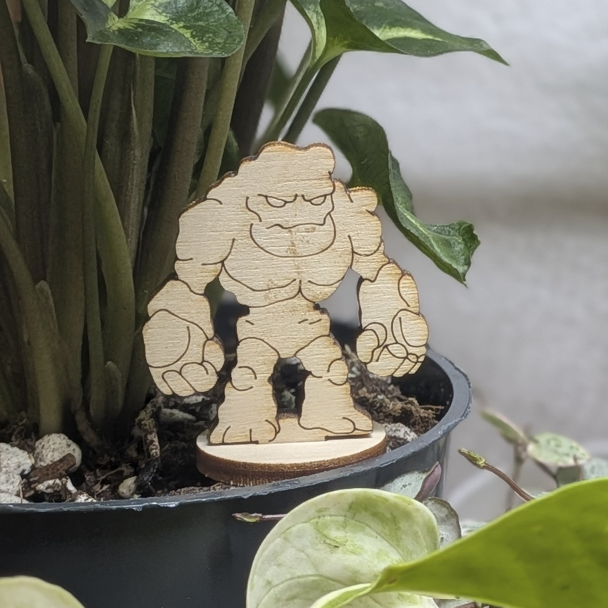 Laser cut plywood stone golem sits in a plant pot. Features jagged rocky body, blank eyes, and natural wood grain texture