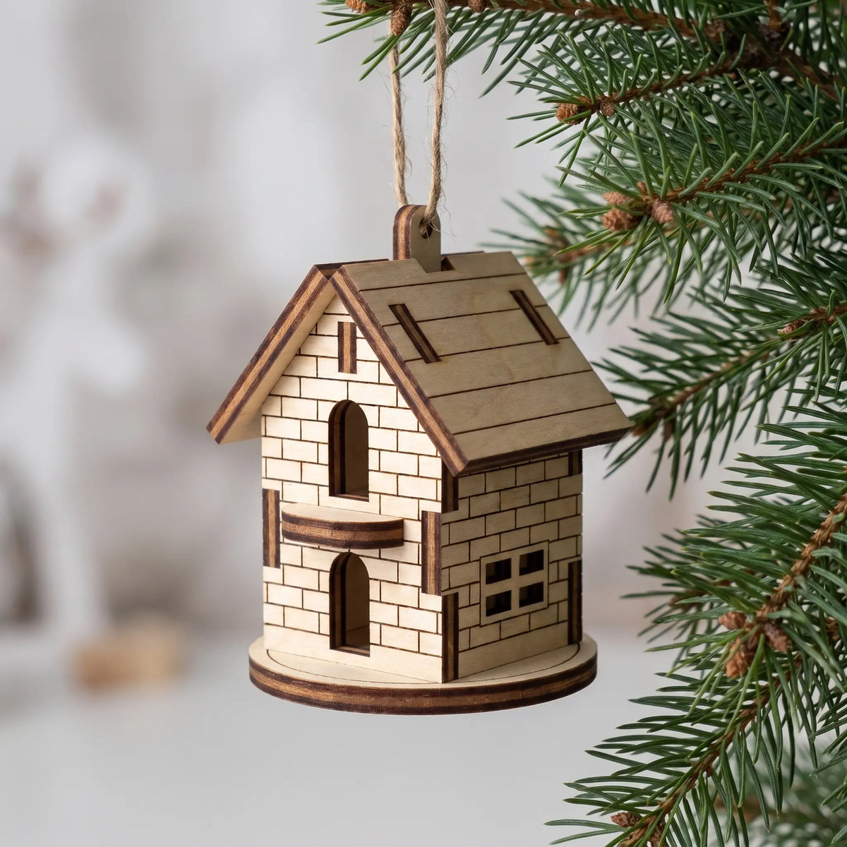 Side view of laser cut plywood Christmas house ornament hanging on spruce tree branch, showing arched door and window panes.