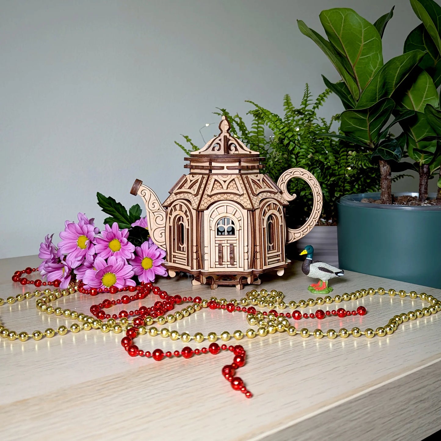 Miniature laser cut plywood teapot model on a table with bright purple flowers, a tiny toy duck, and bead chains.