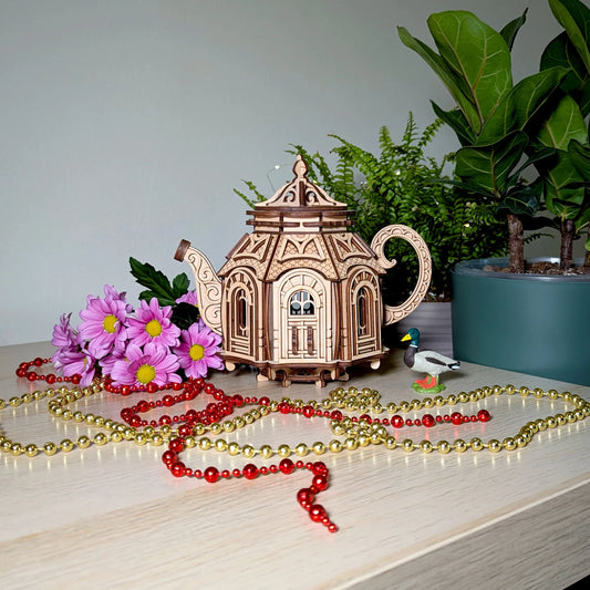 Miniature laser cut plywood teapot model on a table with bright purple flowers, a tiny toy duck, and bead chains.