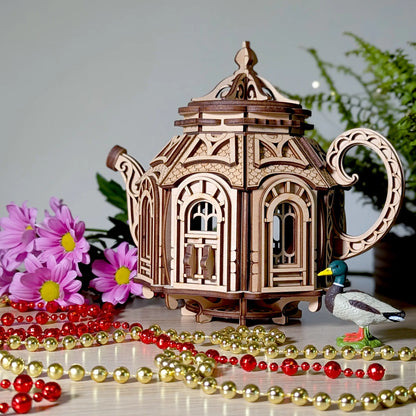 Finished laser cut plywood teapot house staged on a light wood table with a toy duck, flowers, and shiny beads.