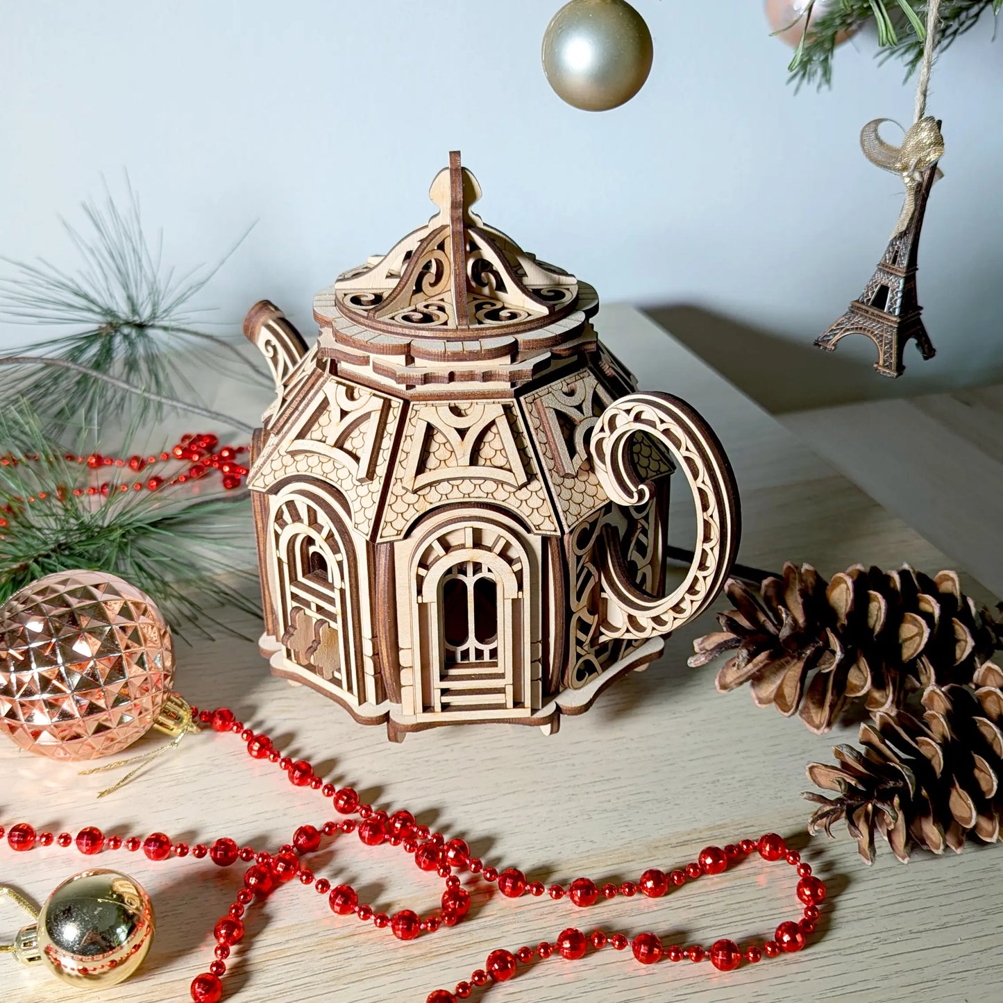 High-angle view of a decorative laser cut plywood teapot house model surrounded by holiday festive ornaments.