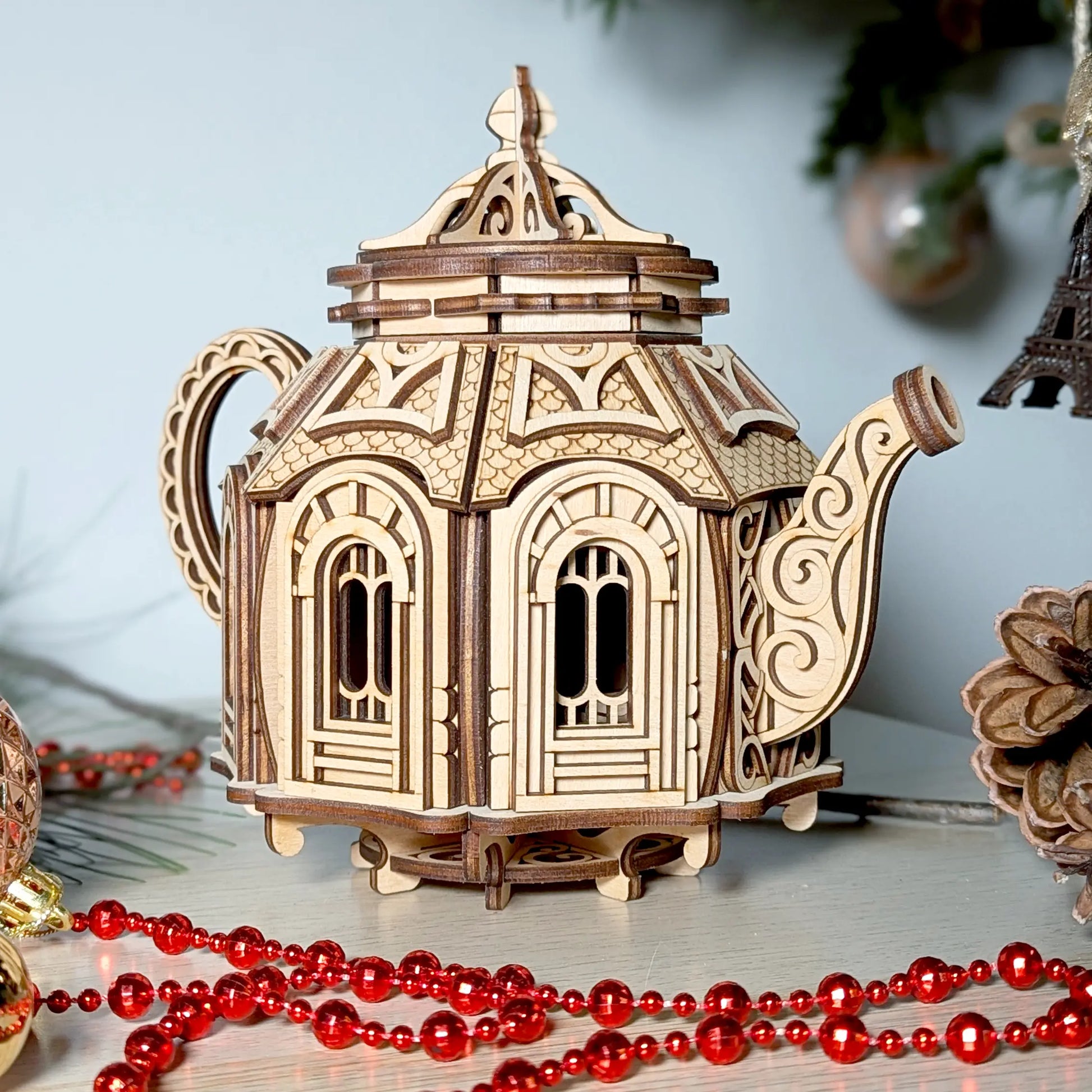 Front view of a miniature laser cut plywood teapot house staged on a table with a festive red beaded garland.