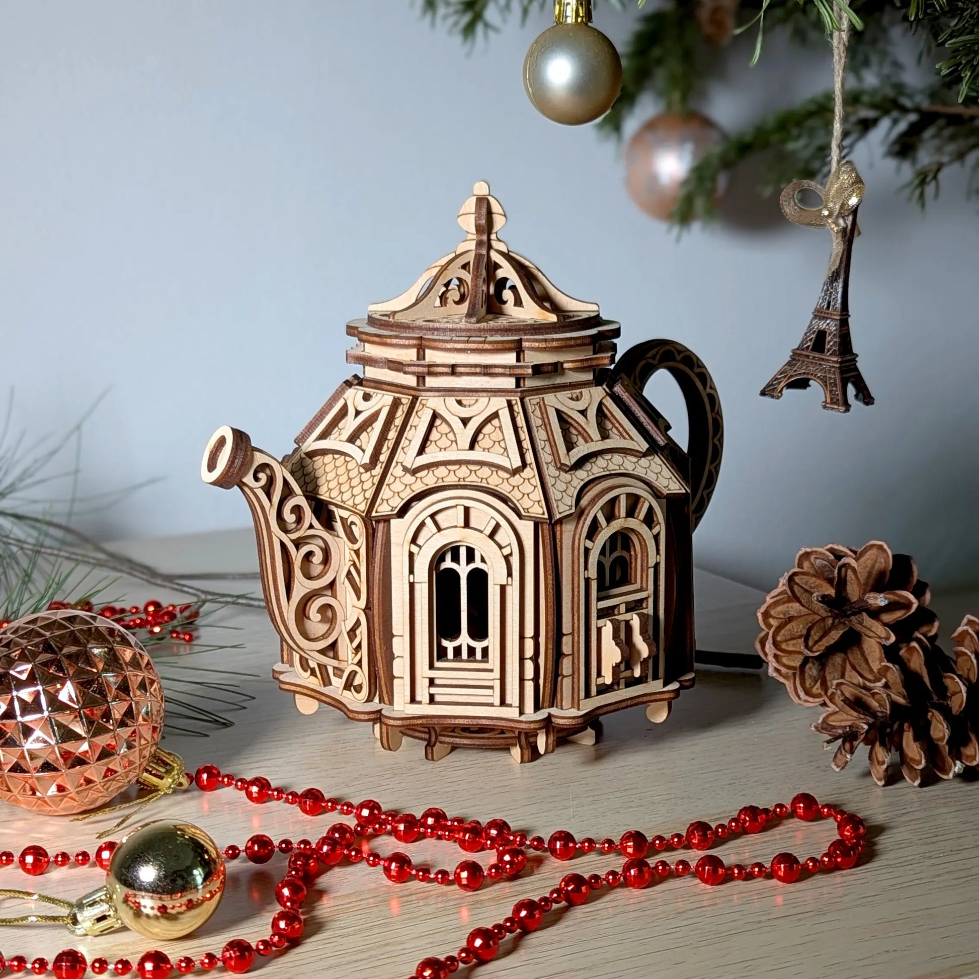 Laser cut plywood teapot fairy house model showcased alongside pine cones and holiday tree decorations.