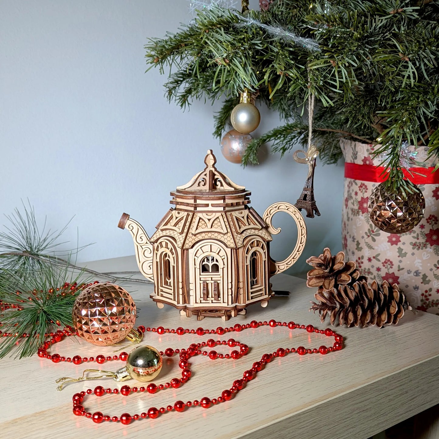 Miniature laser cut plywood teapot fairy house staged beautifully beneath a decorated holiday pine tree setup.