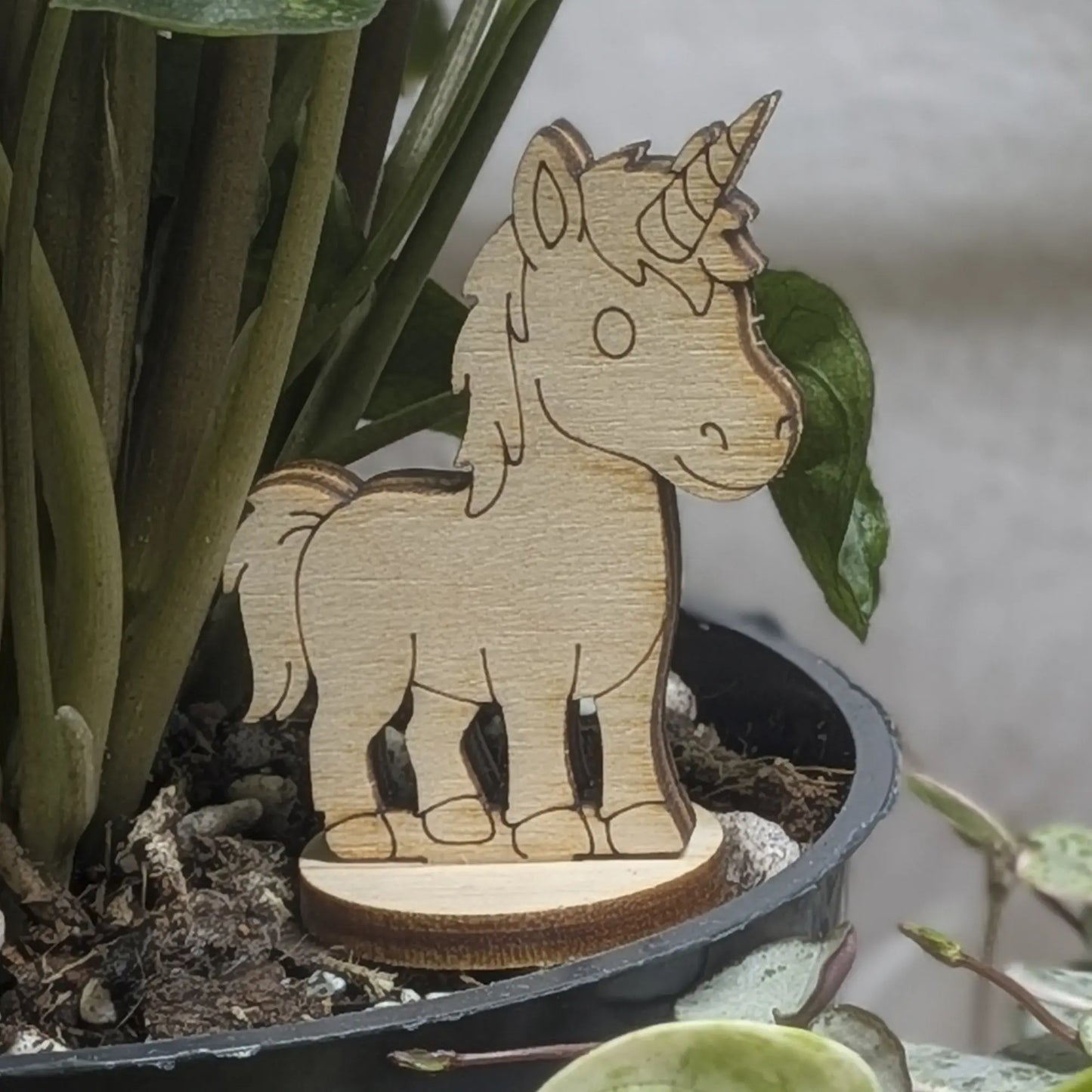 Laser cut plywood unicorn figurine stands in a plant pot. Features engraved spiral horn, mane, and tail on a round base