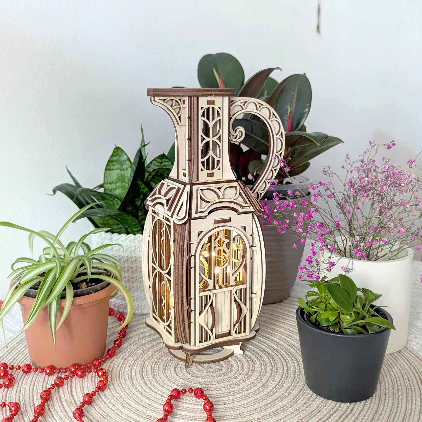 Assembled laser cut plywood jug fairy house glowing with warm internal light on a table surrounded by green houseplants.